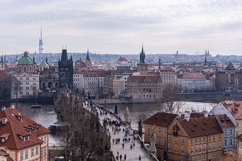 Overlooking Prague's historic skyline featuring Charles Bridge bustling with tourists, with the Vltava River and distant landmarks under a cloudy sky