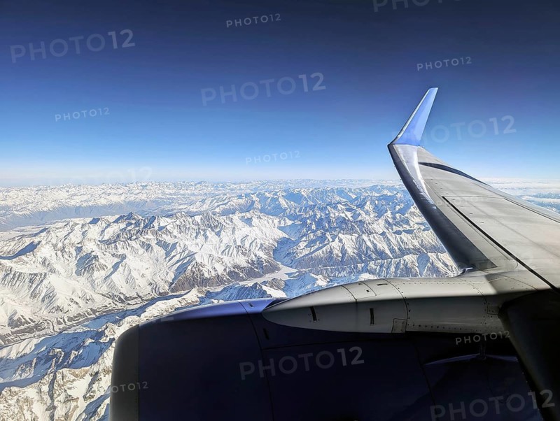 Aerial view of snow-capped mountains seen through an airplane window, showcasing a stunning panorama under a clear, bright blue sky. The airplane wing is visible in the foreground