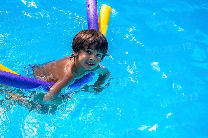 Adorable young child holding colorful pool noodles, swimming and smiling in bright blue water on a sunny summer day, enjoying leisure and children's recreation