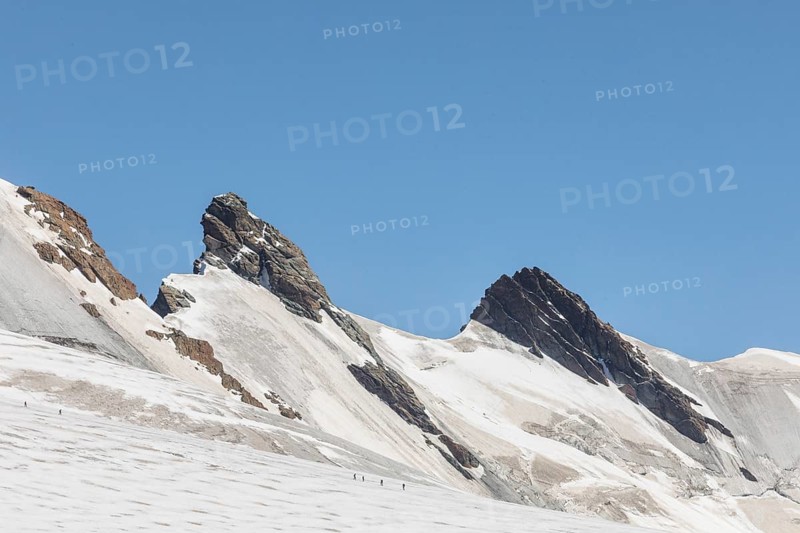 A stunning panoramic view of the Zermatt Alps in summer, showcasing rugged mountain peaks under a clear blue sky with hikers trekking across the glacial plains below