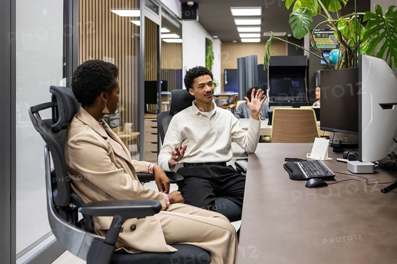 Colleagues engage in a lively discussion at a modern office desk, surrounded by computers and plants. The environment reflects teamwork and professional interaction