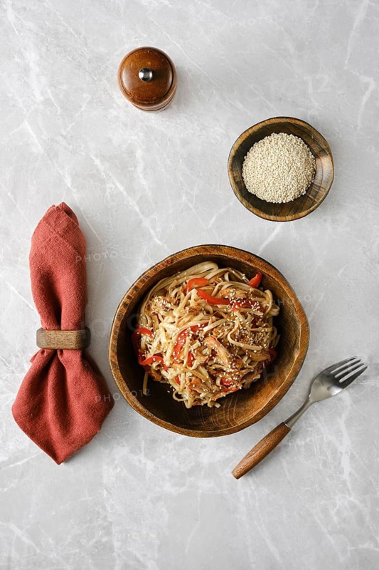 A bowl of stir-fried noodles and pulled beef with red peppers and garnished with sesame seeds rests on a marble countertop. A wooden bowl of sesame seeds and a red napkin accompany it