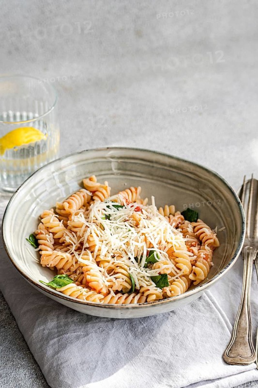 A ceramic bowl filled with cavatappi pasta topped with cheese, fresh spinach and spicy tomato sauce. A glass of water with lemon and silverware are placed beside the dish