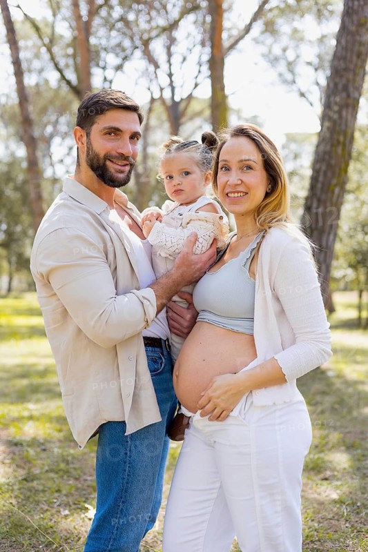A joyful pregnant woman with her family, including a man and a young child, enjoying time outside in a natural setting, surrounded by tall trees and greenery, symbolizing love and togetherness