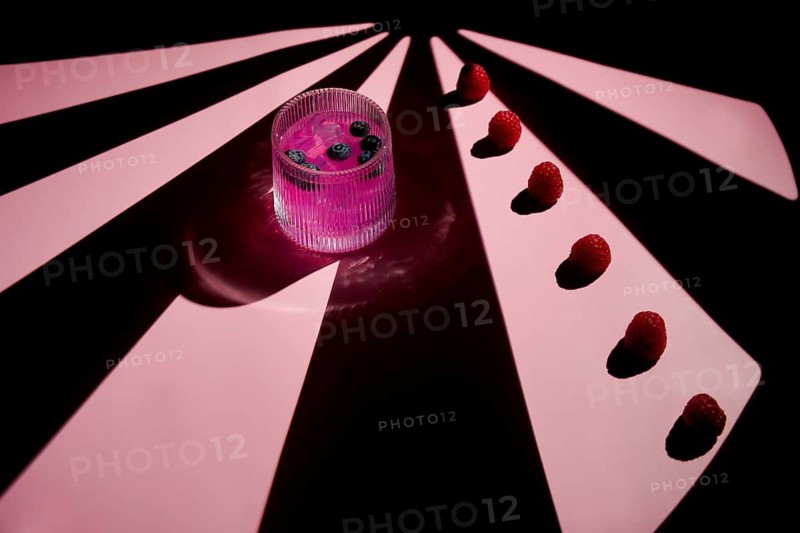A vibrant pink cocktail in a glass, garnished with blueberries, is set against a striking pink-lit background with a pattern of light and shadow. Raspberries are aligned artistically