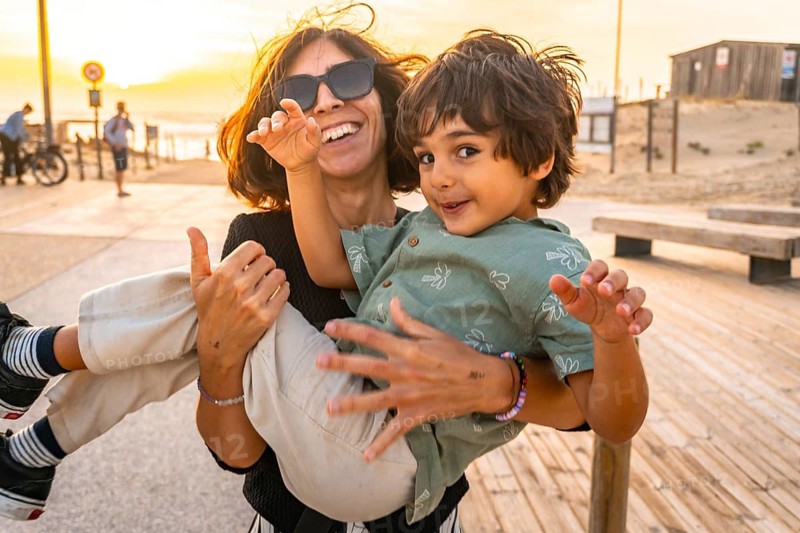 Happy mother holding her young son, both smiling and having fun outdoors on a wooden boardwalk at the beach during a beautiful sunset, sharing a precious family moment