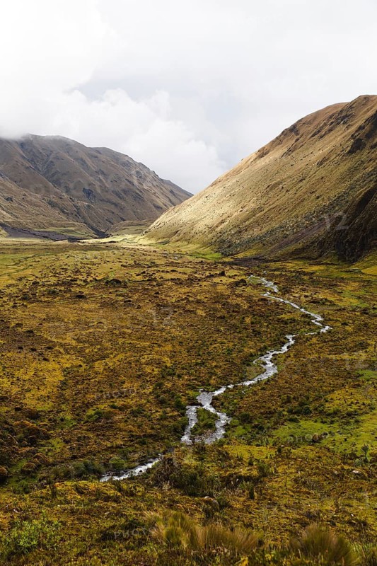 A meandering stream flows through a vast Andean valley at El Altar volcano, Ecuador, surrounded by rolling hills, golden grasses, and dramatic light and shadow contrasts