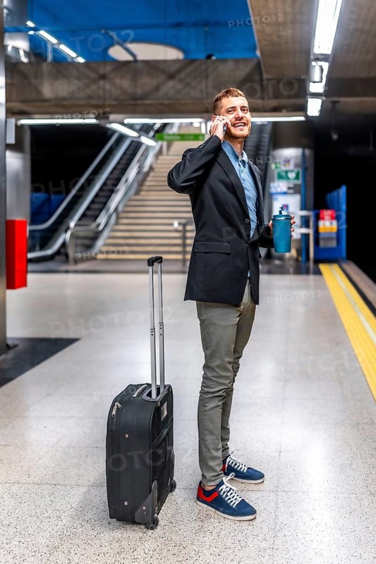 Young businessman talking on phone while waiting for subway train, holding reusable coffee cup and trolley bag