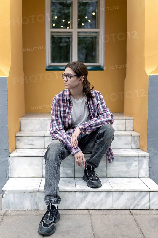 A teenage boy with long hair and glasses sits pensively on marble steps outside a brightly painted building. He wears a plaid shirt, jeans, and sneakers, looking thoughtful