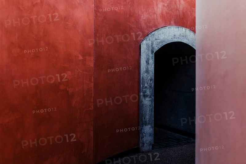 This image features a minimalist architectural design with a striking contrast between vibrant red walls and a weathered gray arched doorway. The simplicity and bold color use highlight the essence of modern minimalism