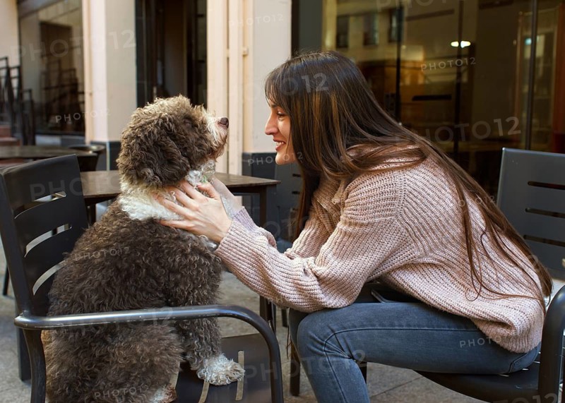 A young woman with long hair enjoys a moment with her dog at an outdoor cafe. She lovingly holds the dog, conveying warmth and companionship in a casual setting