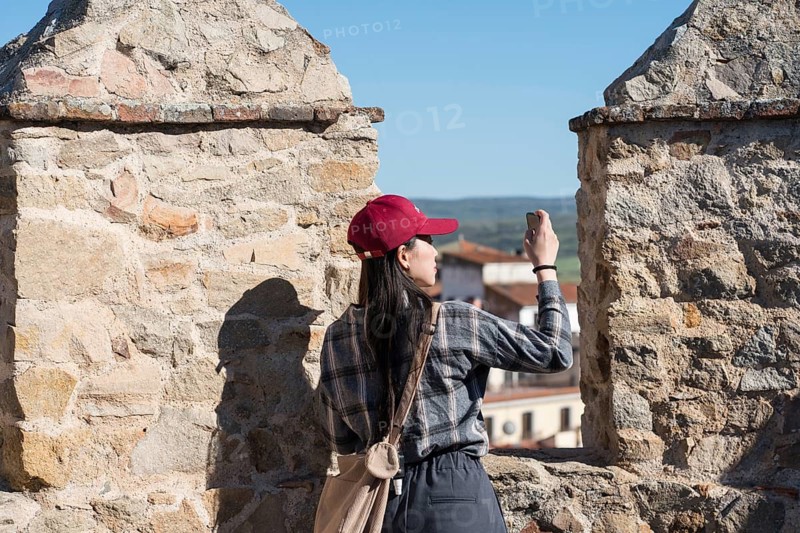 An Asian woman captures photos while standing on the medieval walls of Avila, Spain, with scenic views and ancient stonework, highlighting her travel experience