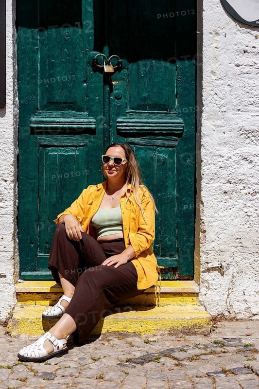 A woman tourist enjoys a sunny day sitting on steps in a charming street. Dressed in vibrant colors, she exudes a laid-back vibe, embracing her travel experience
