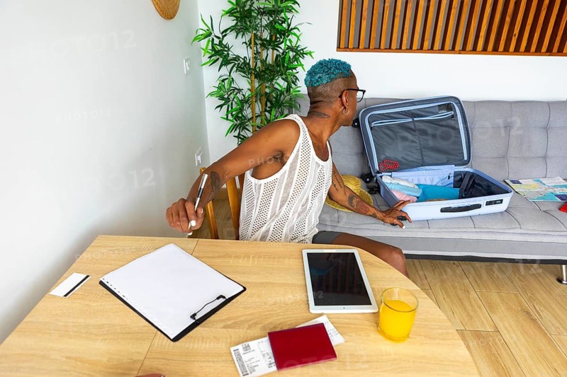 An LGBTQIA+ man with blue hair and a white top organizes travel documents and a suitcase in a cozy living room setting, preparing for the holidays
