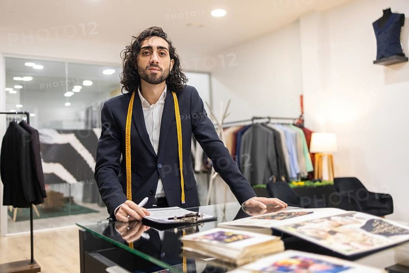A professional tailor in a sleek boutique leans on a glass table surrounded by measuring tape and design books. Racks of suits and cozy lighting create an elegant atmosphere