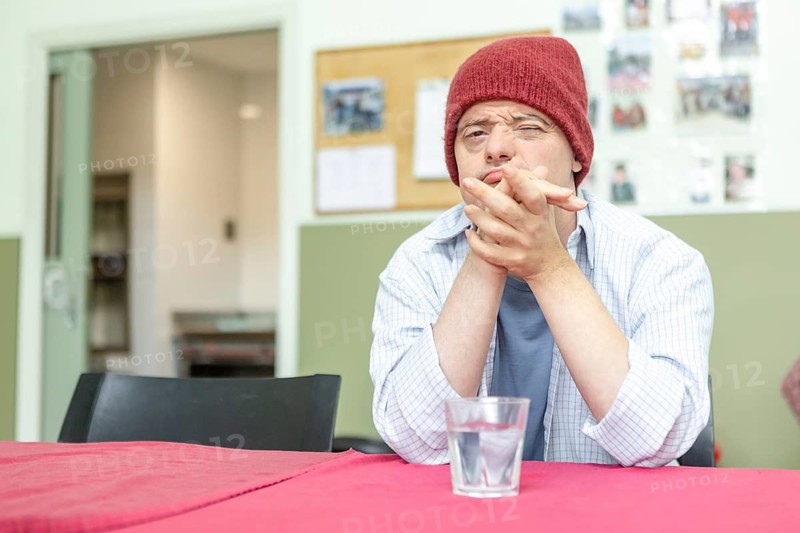 A man in a mental health residence sits contemplatively at a table, wearing a red beanie and casual attire. A glass of water is in front of them, enhancing the serene atmosphere