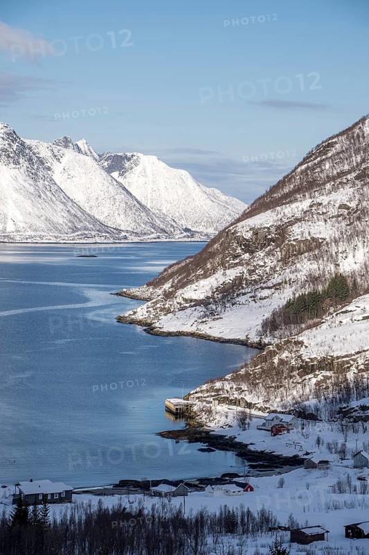Scenic view of Mefjordbotn fjord, Norway, showcasing snow-covered peaks and a serene blue sea under a clear sky, with cozy houses dotting the snowy landscape below