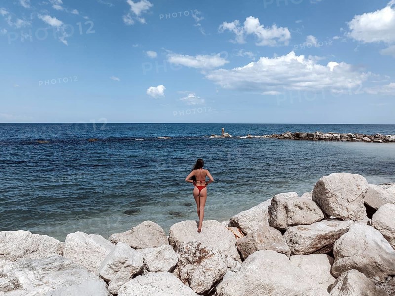 A young woman stands on the rocky shores of an Italian seaside, admiring the vast expanse of the blue ocean under a clear sky. The scene captures the essence of tranquility and nature