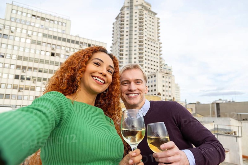 Two cheerful friends pose for a selfie while holding glasses of white wine on a rooftop The city skyline in the background enhances the vibrant and fun urban atmosphere