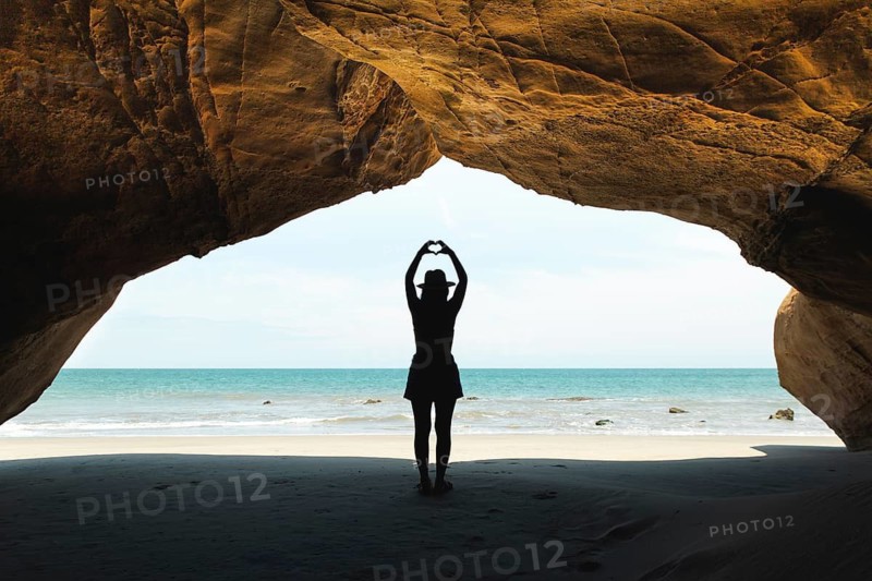 A young Latin woman stands under a rocky arch in Mancora, Peru, forming a heart shape with her hands. The serene beach and ocean vistas frame her silhouette