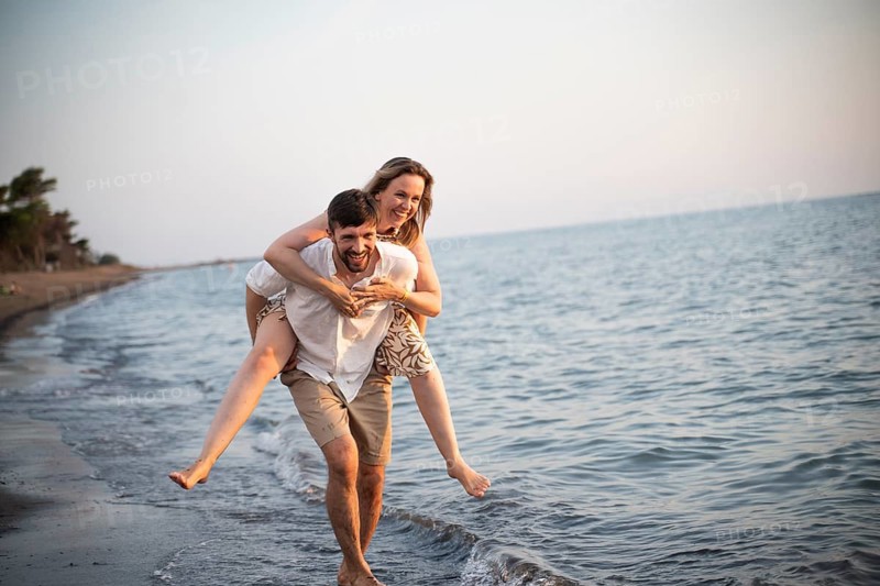 A joyful couple enjoys a playful day at an Italian beach, with one partner giving the other a piggyback ride. The serene sea and soft sand create a perfect vacation scene