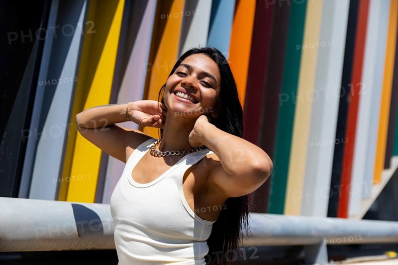 A woman in a vibrant outfit poses against modern architectural elements. The colorful backdrop creates a striking urban contrast with her joyful expression