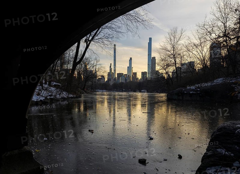 A serene sunset illuminates the New York skyline, seen across a frozen pond from under a bridge in Central Park. Late winter scenery provides a calm yet stark contrast between nature and urban life