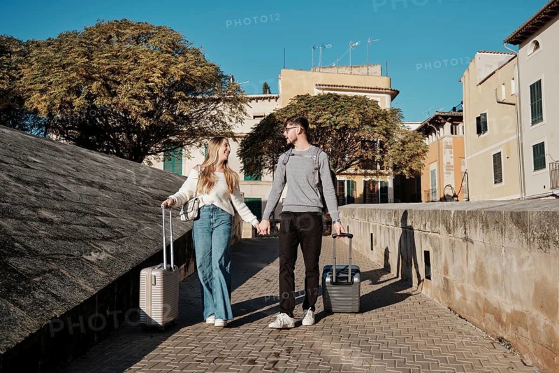 A young couple walks hand in hand with suitcases, exploring the charming streets of Majorca, Spain, as they search for their ideal vacation home. Sunny and adventurous vibe