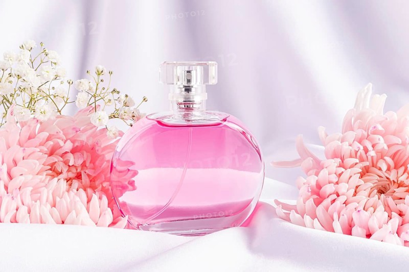 A round glass perfume bottle filled with pink liquid, surrounded by delicate pink and white flowers on a soft white fabric, creating a sophisticated and elegant scene