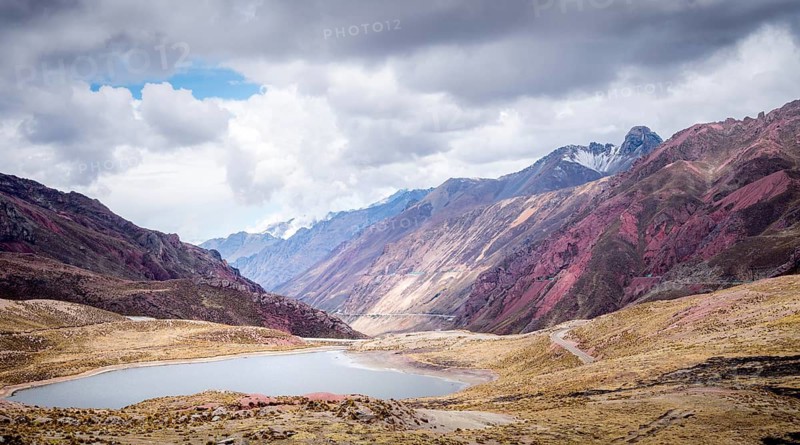 Majestic red mountains of the Peruvian Andes surround a tranquil lake under dramatic skies. A breathtaking blend of vibrant hues and natural beauty in a serene landscape