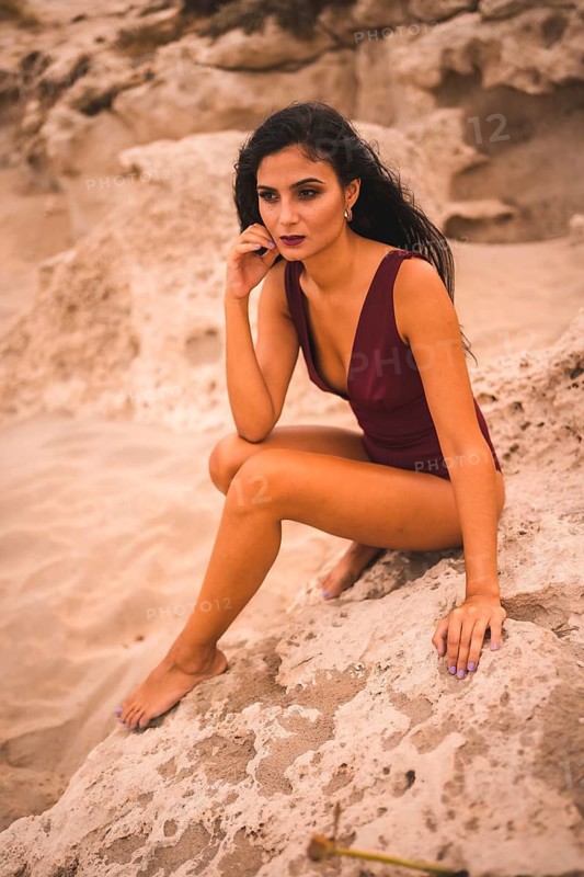 Caucasian brunette in a maroon swimsuit, sitting smiling on the beach of Cabo de Gata, Nijar. Andalucia, Spain