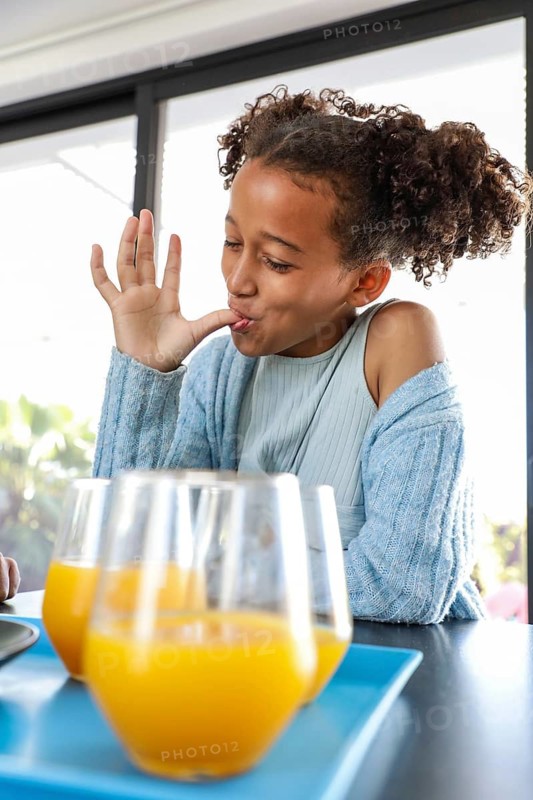 A young girl in a cozy sweater enjoys a tasty snack while savoring the moment, with three glasses of fresh orange juice on the table in a sunlit room