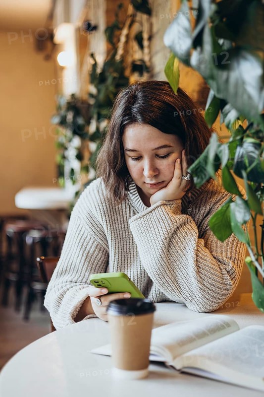 A young woman in a sweater reads a book at a table in a bookstore Surrounded by plants, she holds a phone and drinks coffee, creating a cozy, relaxed atmosphere