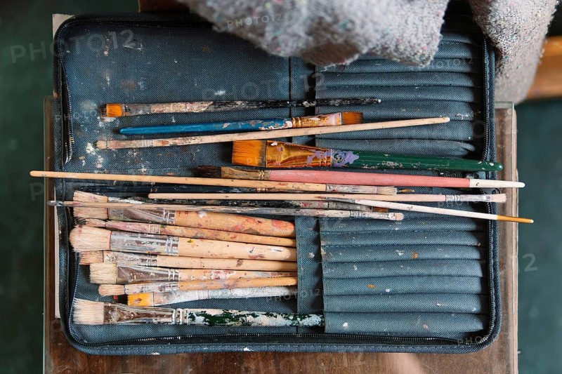A set of well-worn paintbrushes, covered in dried paint, rests in a fabric case on an artist's table. The assortment of brushes reflects the creativity and passion of a painter