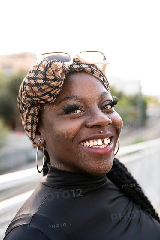 A cheerful young african woman with a patterned headscarf and stylish hoop earrings smiles warmly outdoors, showcasing her vibrant personality and radiant energy