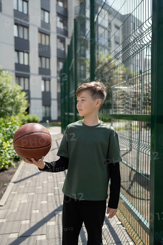 A young boy stands on a paved path in an urban setting, holding a basketball in one hand. He is dressed casually and appears focused on his surroundings, near a sports area