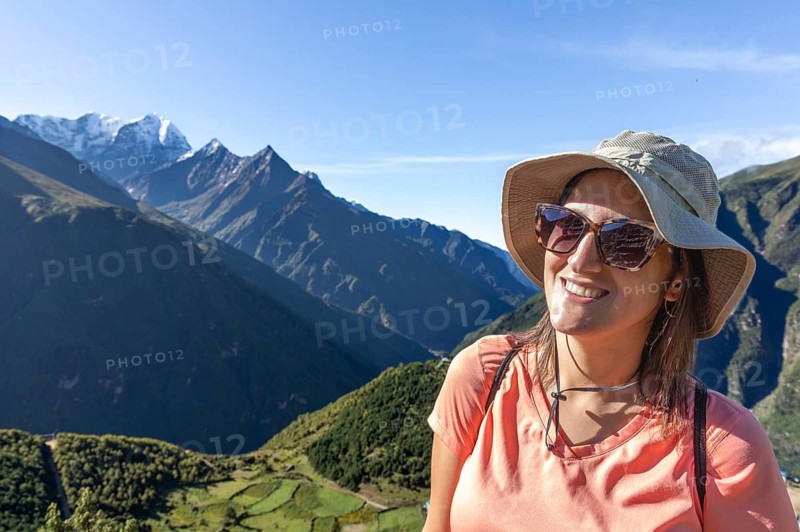 Smiling woman wearing a hat and sunglasses enjoys a scenic trek in the Everest region, with stunning mountain peaks and clear skies in the background, capturing the spirit of adventure