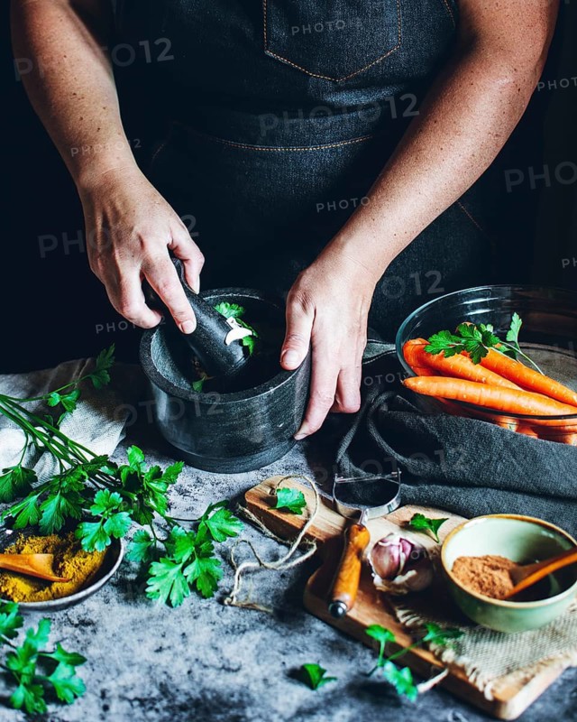 Hands crush fresh herbs in a mortar, surrounded by vibrant vegetables and spices. The scene captures the essence of traditional cooking with rustic ingredients and tools