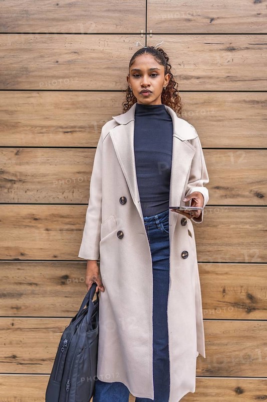 Vertical portrait of a young african american businesswoman in coat holding mobile and laptop bag outdoors leaning on a wooden wall