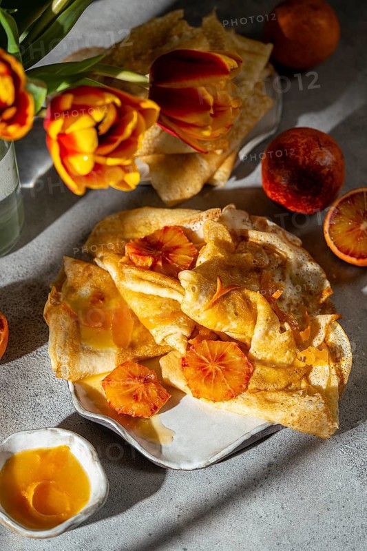 A delightful presentation of Crepes Suzette, featuring thin pancakes draped in a rich, caramelized blood orange sauce, accompanied by fresh blood orange slices and tulips