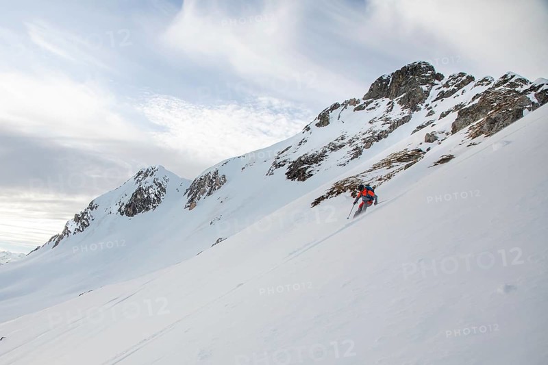 A skier descends a steep snowy slope under a partly cloudy sky. This image captures the essence of backcountry skiing, highlighting the adventure and natural beauty