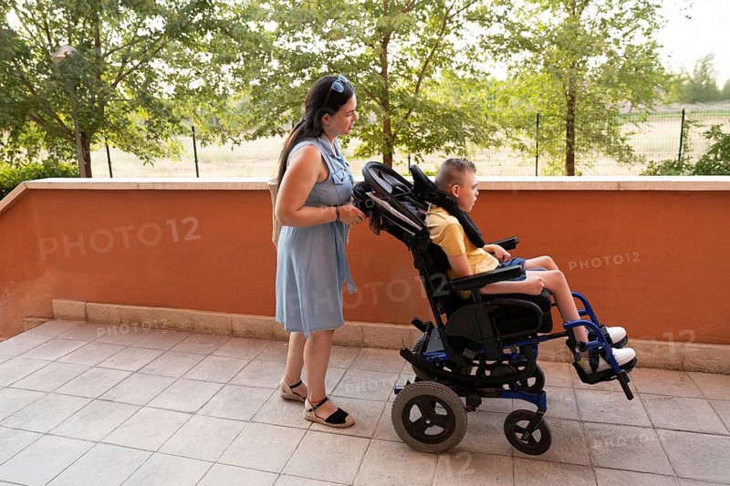 A mother attentively assists her child in a wheelchair on a sunny terrace, showcasing care and support. The vibrant setting highlights their positive interaction and bond