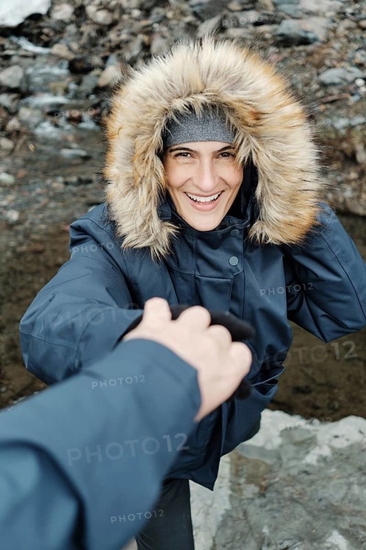 A joyful woman in a warm winter jacket with a fur-lined hood reaches for a helping hand while exploring a rocky, snowy landscape