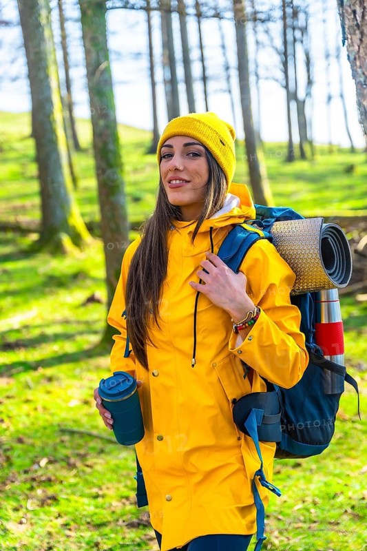 Hiker woman with yellow raincoat and backpack walking in forest, enjoying trekking and drinking hot beverage