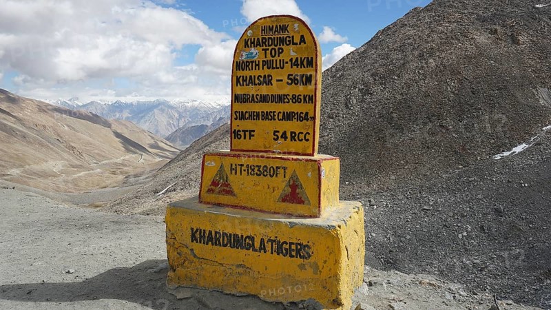 Signpost on dusty Khardung-la Pass in the mountains with sweeping views of surrounding mountains and cloudy skies, trekking in Ladakh, Himalayas, India