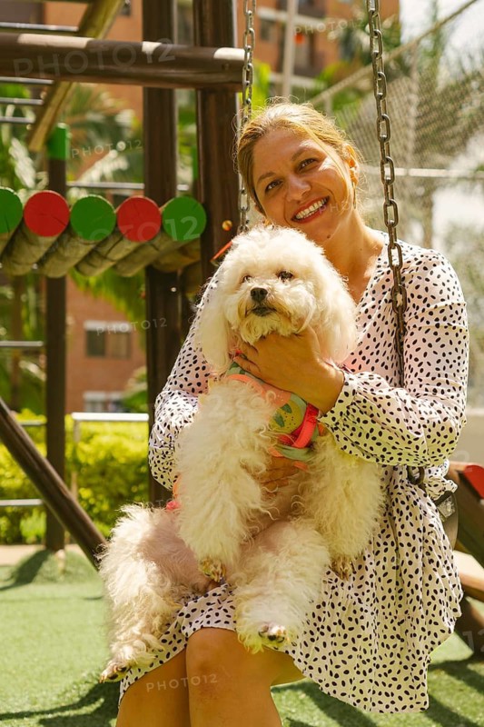 Smiling woman sitting on a swing in a vibrant playground, joyfully hugging her fluffy white poodle in a pink harness, relishing the warmth of a sunny day outdoors