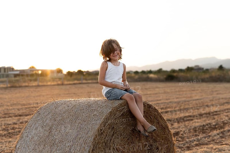 A young child sits on a hay bale in a field at sunset, enjoying the serene rural landscape. The golden hour light casts a warm glow over the peaceful scene
