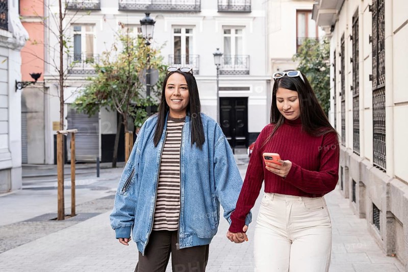 A Latin lesbian couple walks hand in hand down a charming urban street. They are smiling and engaged in conversation, highlighting their connection and joy