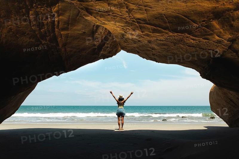 A young woman stands in a cave with arms raised, facing the ocean at Mancora, Peru. The image captures the essence of freedom and adventure by the sea