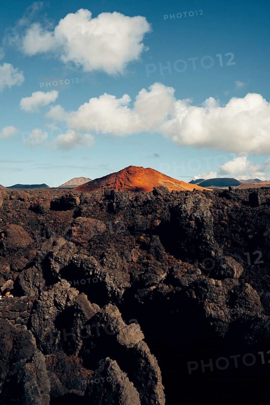 Stunning view of Lanzarote's volcanic terrain, showcasing dramatic rock formations with vibrant red and black hues, under a clear blue sky dotted with fluffy clouds